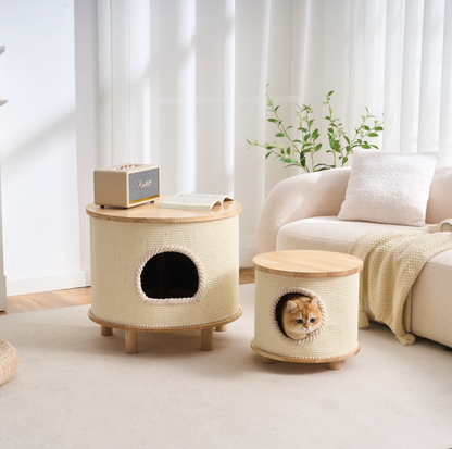 Two round cat houses in a warm cream woven fabric with light wood tops and legs, shown in a bright living room. One cat house has a round entrance opening and a small speaker on top, and the other has a circular opening with a ginger cat peeking out.